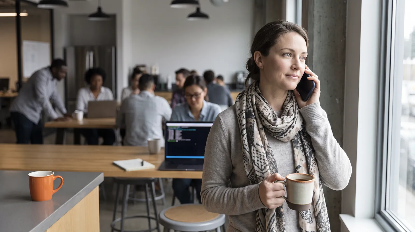A developer by the office kitchen window, talking softly into her phone with a coffee in her other hand, her laptop at the desk behind her.